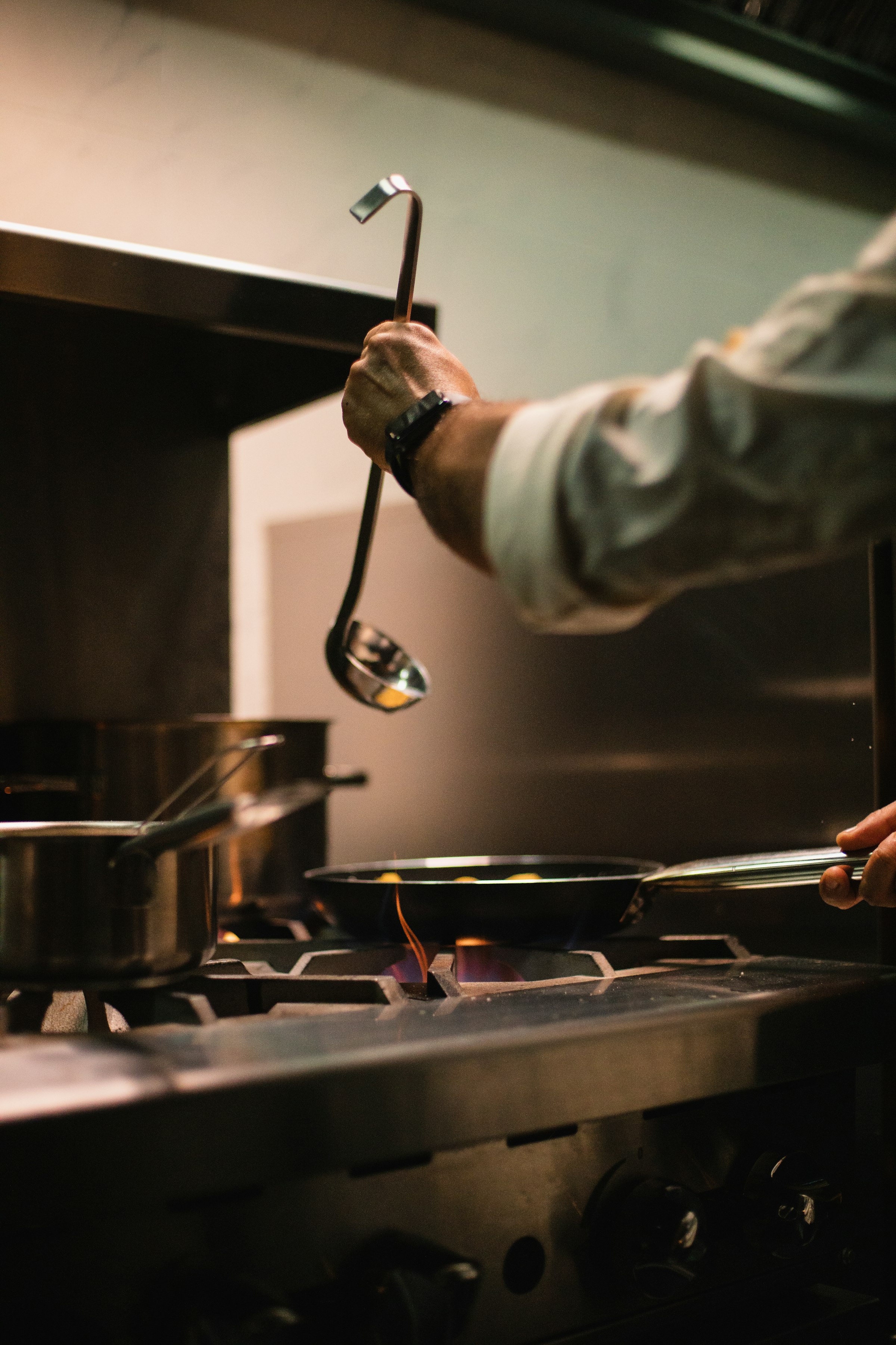 A chef's hand holding a ladle over a pan on an open flame in a dark kitchen