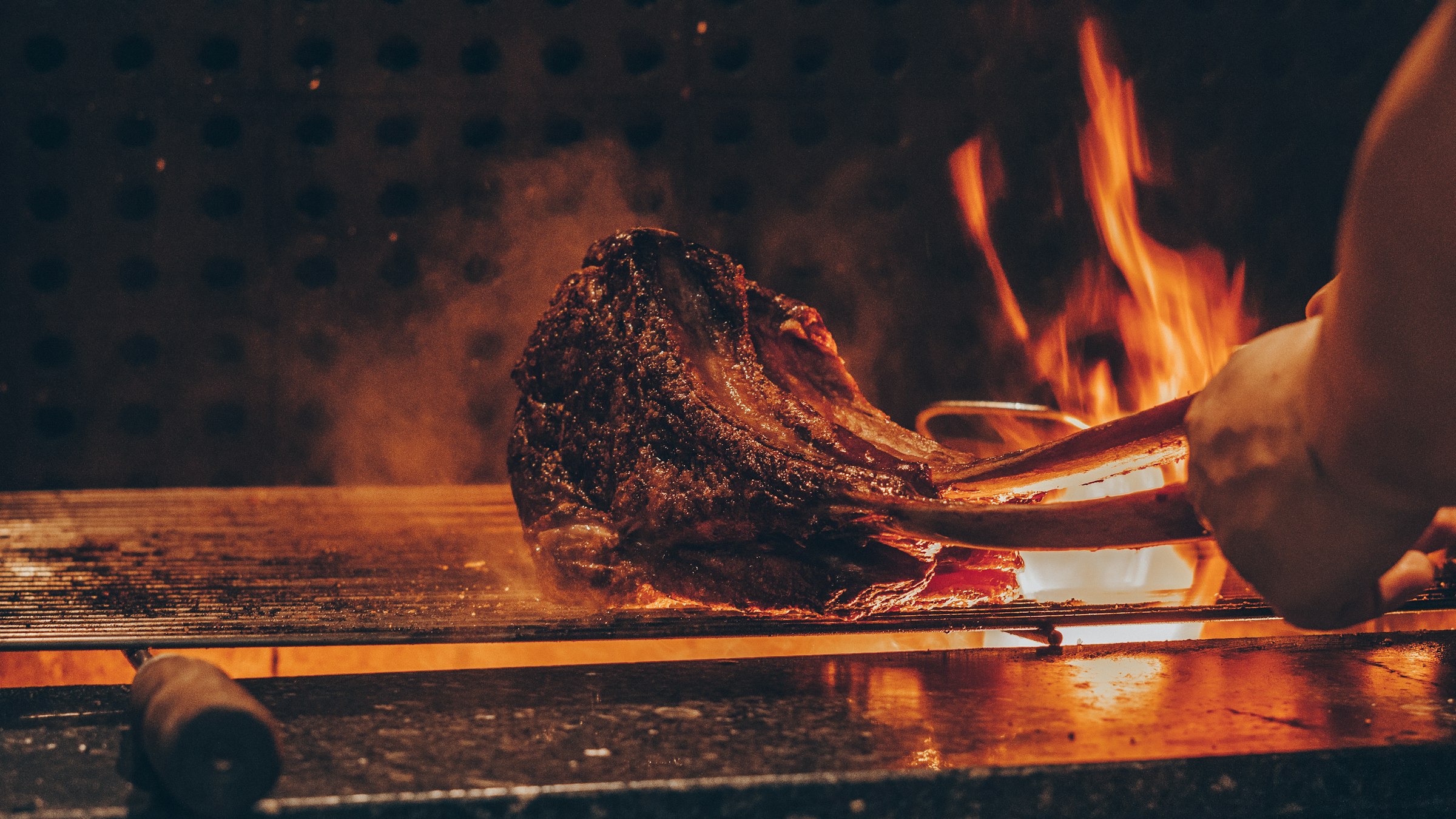 A dry-aged tomahawk steak searing over open charcoal flame, tongs lifting it from the grill